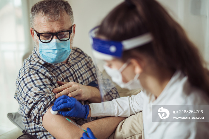 Young woman nurse with surgical mask and face shild giving injection to senior man at home or in a nursing home. Covid-19 or coronavirus vaccine