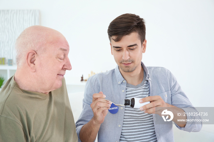 Young male volunteer giving medicine to senior man in light room