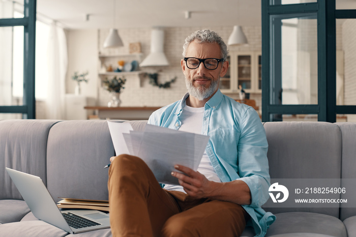 Busy senior man checking the papers and using laptop while sitting on the sofa at home