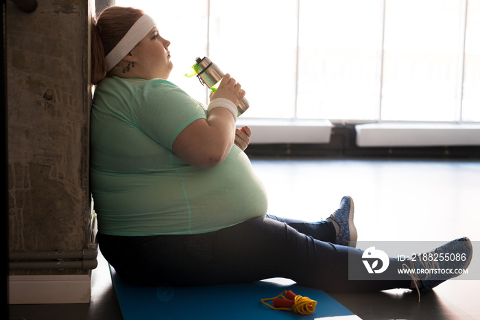 Full length side view portrait of pensive fat woman sitting on mat and drinking water while taking break in workout