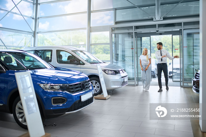 Competent salesman walking with female client at modern auto salon and presenting various luxury car. Young caucasian woman choosing new vehicle at showroom.