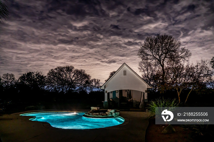 A pool, spa and cabana house at night under cloudy sky illuminated from underneath by a distance rising sun, Hill Country, Texas