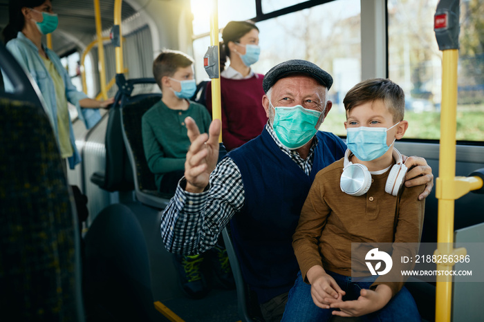 Happy grandfather and grandson talking while wearing face masks and commuting by bus together.