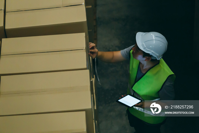 Top view of young warehouse worker man with safety hard hat is checking order details with a digital tablet at inventory room