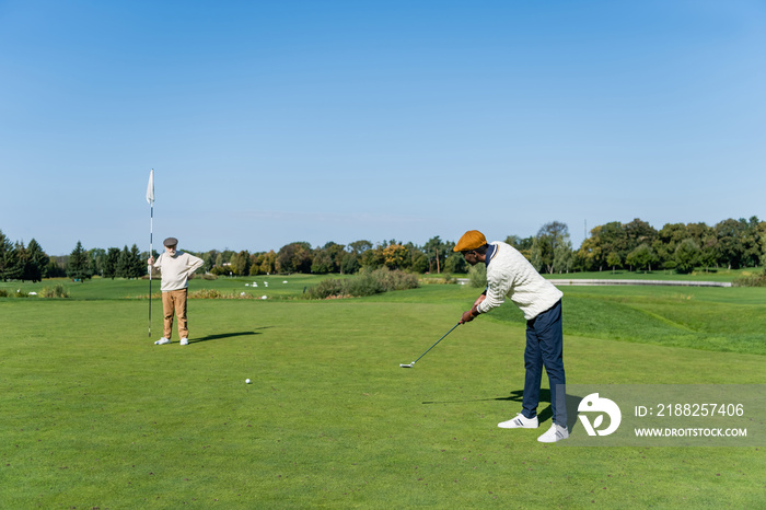 senior man in flat cap holding flag stick while african american friend playing golf.