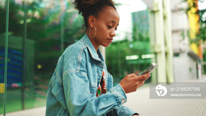 Cute African girl with ponytail, wearing denim jacket, in crop top with national pattern, sitting at the bus stop and using her mobile phone.