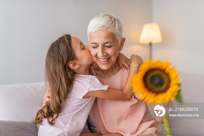 Happy grandmother hugging small cute grandchild thanking for flowers presented, excited granny embrace granddaughter congratulating her with birthday, making surprise presenting bouquet