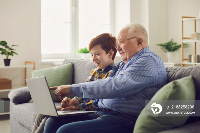 Happy family using modern technology. Smiling grandfather and grandson sitting with laptop computer on cozy sofa at home, enjoying good wifi connection, browsing Internet and watching movies together