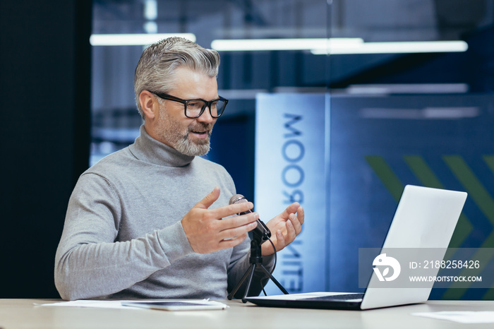 Mature smiling businessman recording audio podcast about finance and strategy to achieve all and good achievement, gray haired man inside office using professional microphone and laptop.