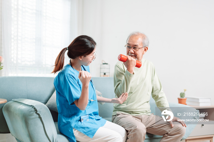 Asian Senior old man doing physiotherapist with support from caregiver. Mature male sitting on sofa in living room using dumbbell workout exercise. Healthcare medical concept.