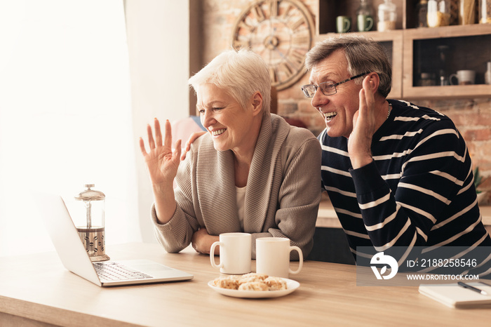 Happy senior couple looking at laptop screen in kitchen and waving