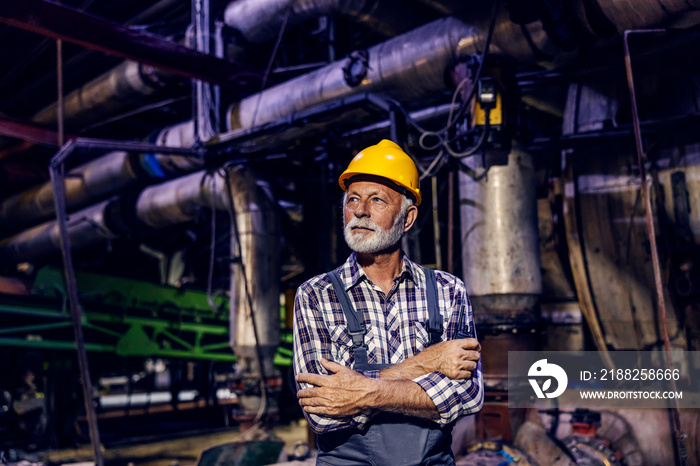 A successful senior factory worker with a helmet on his head is standing in the factory with arms crossed and looking away. Old factory worker looking for new challenges.