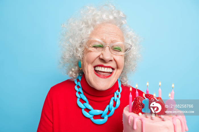 Positive aged woman with grey curly hair smiles broadly has white perfect teeth holds cake celebrates birthday has retirement age isolated over blue background. Grandmother enjoys bday party