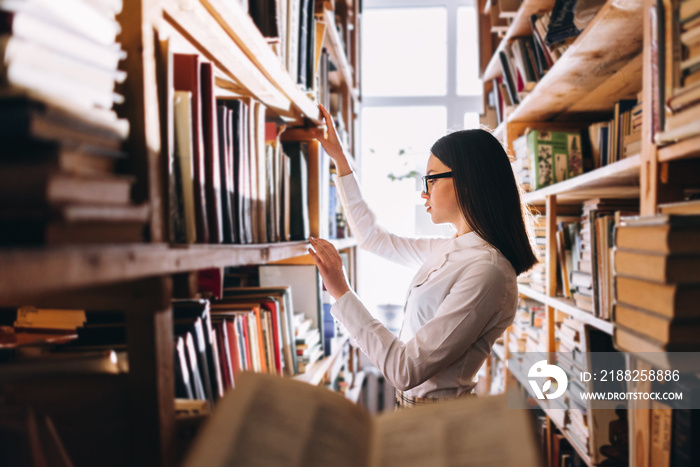 people, knowledge, education and school concept - student girl select a book on the shelf in the old library