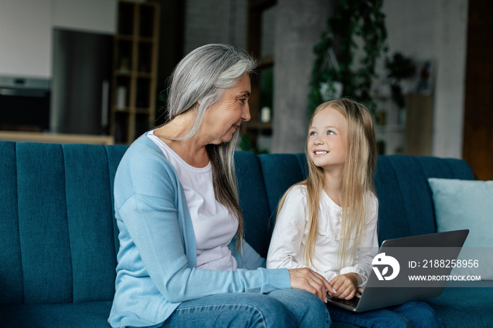 Smiling caucasian small granddaughter and old grandma have video call on computer, study online, play on sofa