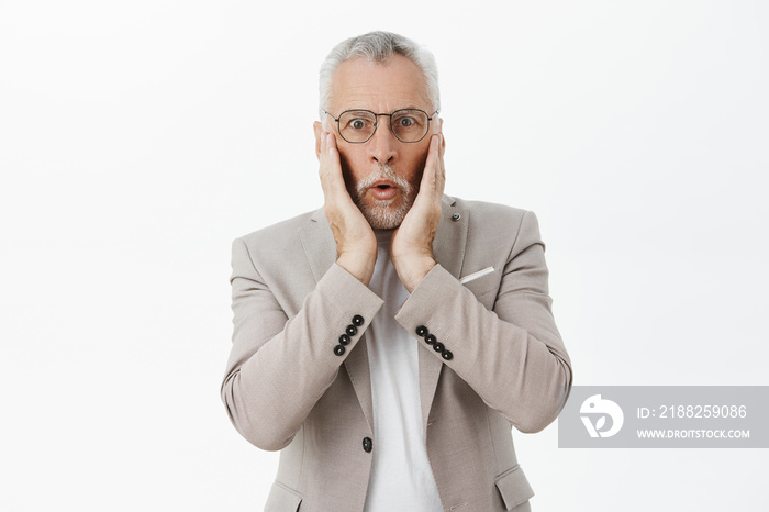 Waist-up shot of shocked impressed handsome senior male entrepreneur in glasses and grey suit pressing hands to cheeks amazed and speechless staring at camera focused from hearing shocking news