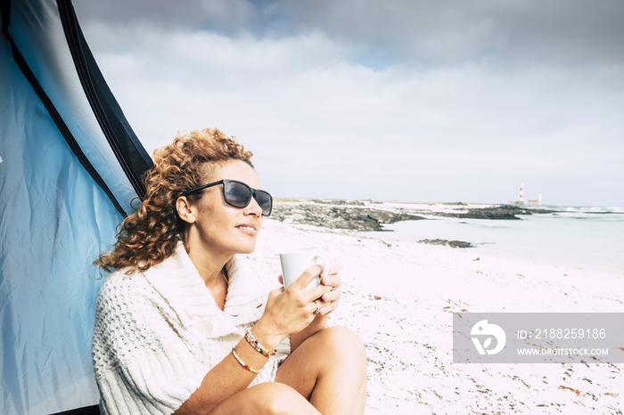 Beautiful middle age woman sit down outside a tent in free beach camping outdoor feeling the nature while drink a tea or coffee from a white cup - wind in her hair and freedom concept for lifestyle