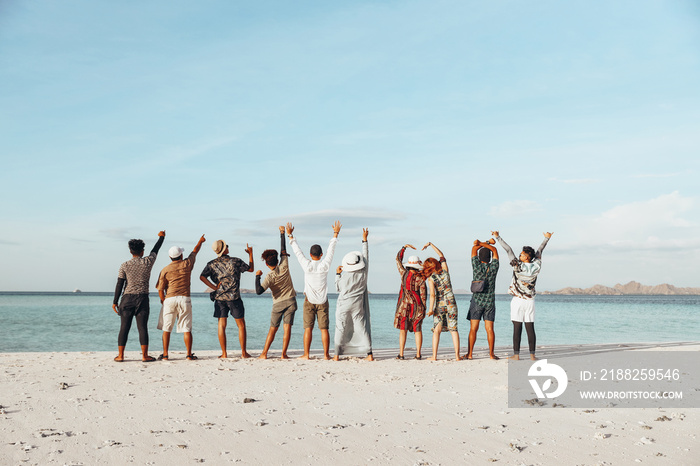 Back view of group of friends standing and raising hand together enjoying the sea view from beach during summer vacation