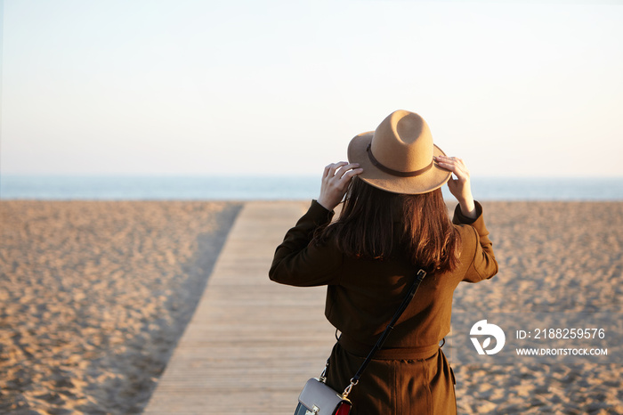 Rear view of unrecognizable brunette girl wearing hat, coat and shoulder bag standing on boardwalk along beach, enjoying nice warm day, came to the sea to make her mind after hard working day