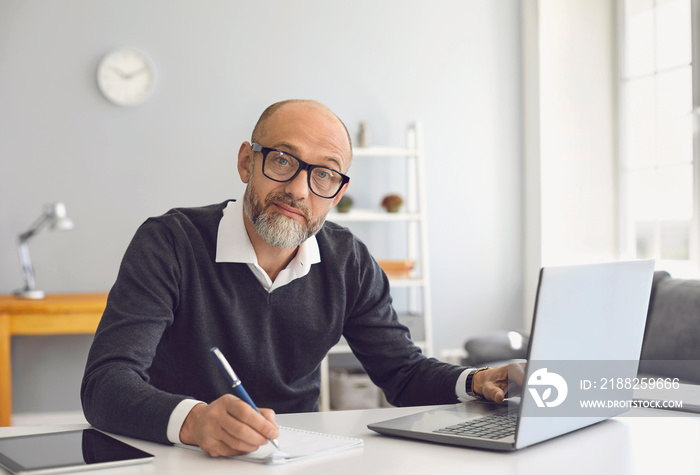 An elderly man with glasses is working he is studying he has a laptop he writes notes in a notebook while sitting at a table in his home office.