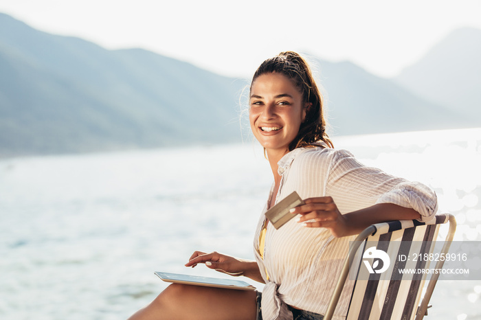 Smiling woman sitting on deck chair by the sea using tablet and credit card on a sunny day