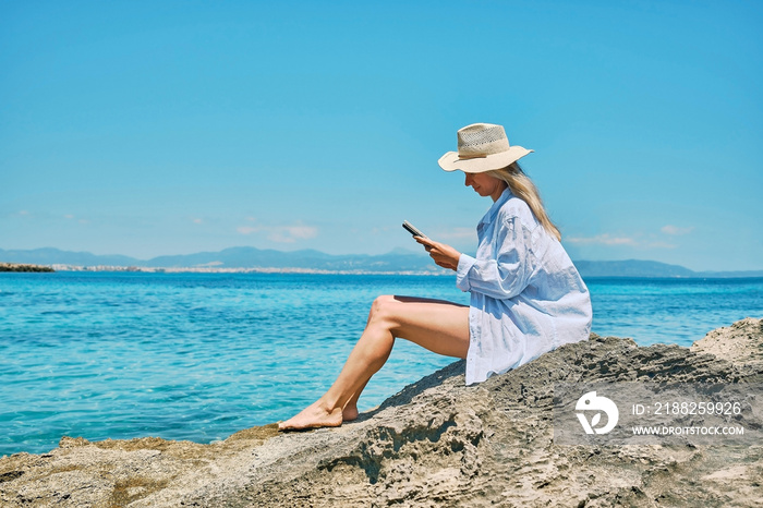 Woman outdoors lifestyle watching, reading on tablet ebook on the beach in summer day. Wearing wide brimmed hat, Sunbating with uv protection. Concept of beach vacation.