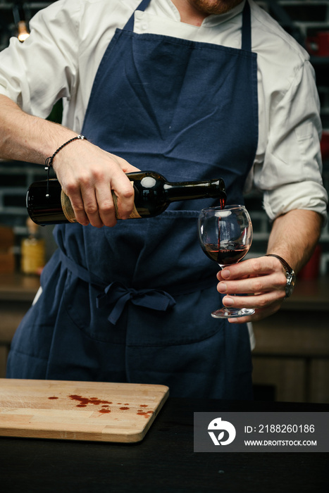 a man in a blue apron and white shirt is standing in the kitchen and opens a bottle of red wine with a corkscrew