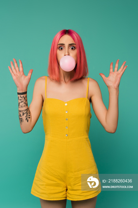 shocked young woman with pink dyed hair blowing bubble gum isolated on blue