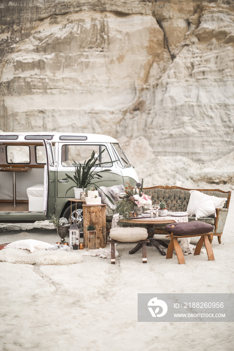 Green retro bus and rustic decor in the canyon. Table in the boho style with candles, succulents, plant decoration, green vintage sofa and chairs with pillows and fur, wedding rustic cake