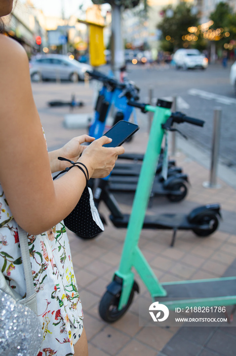 Woman’s hands close up holding a phone near the modern city electric kick scooter, going to unlock it. Modern city transportation.