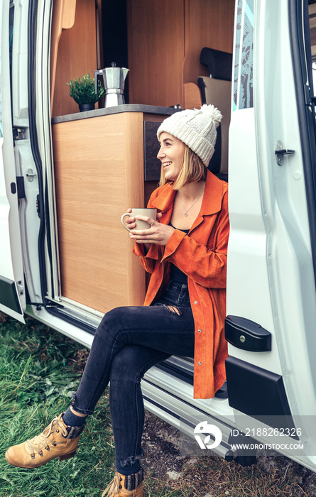 Smiling woman drinking coffee sitting at the door of a camper van