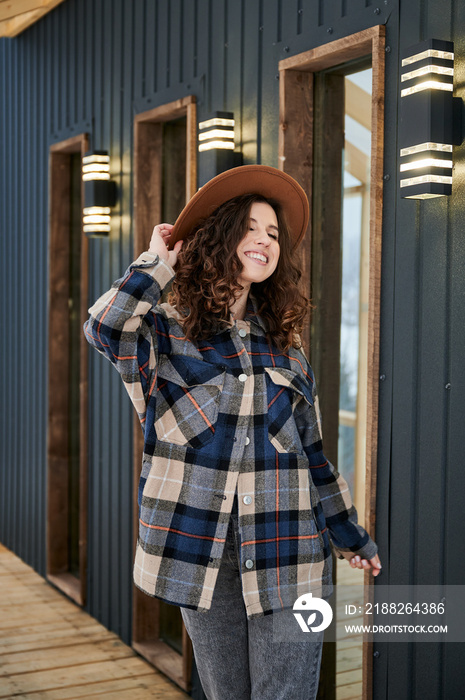 Curly young woman resting on terrace of modern barn house in the mountains. Happy female tourist enjoying winter holidays in new cottage.