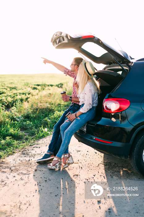 Happy couple hugging at open trunk of hatchback car and pointing finger outdoors