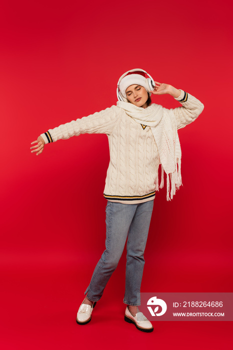 Young woman in santa hat and knitted clothes listening music in headphones on red background.