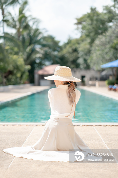 Woman wearing white shirt, long skirt and straw hat posing near swimming pool.