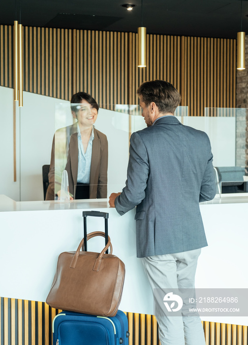 Female Hotel Receptionist Assisting Businessman for Checking In. Business man with luggage talking with concierge on hotel reception with sneeze guard protection.