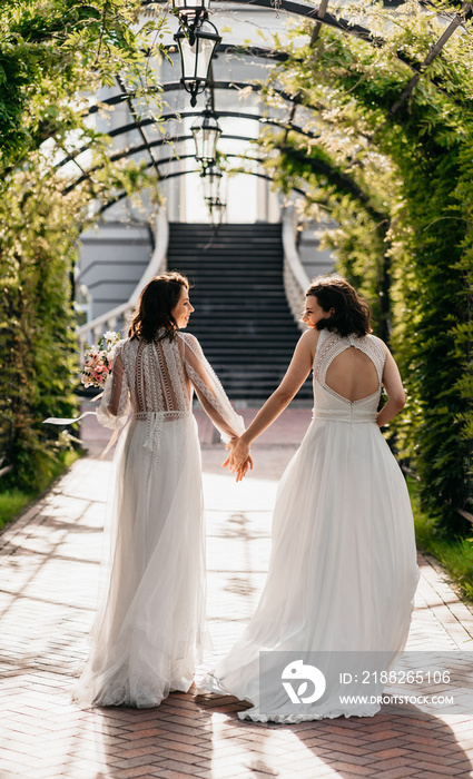 Two happy lesbian LGBT brides wearing boho dresses walking through alley on their wedding day