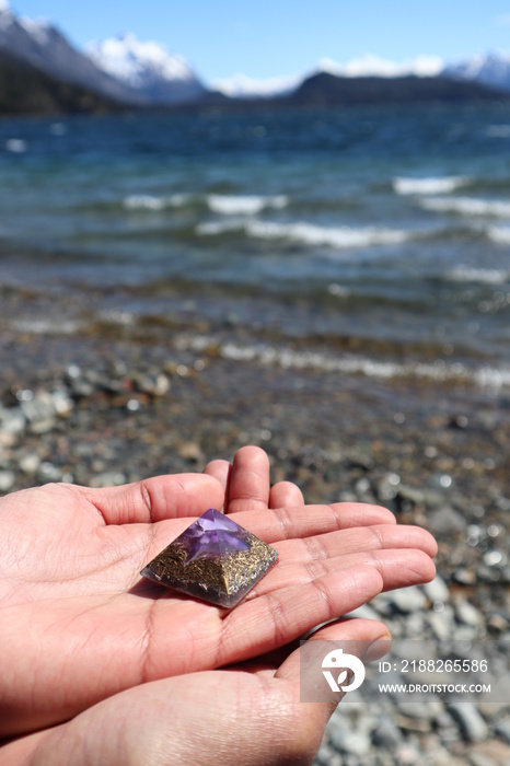 Orgone structure over a stone in a lake shore with mountains in the background. Hands are holding an orgone stone. Natural healing power.