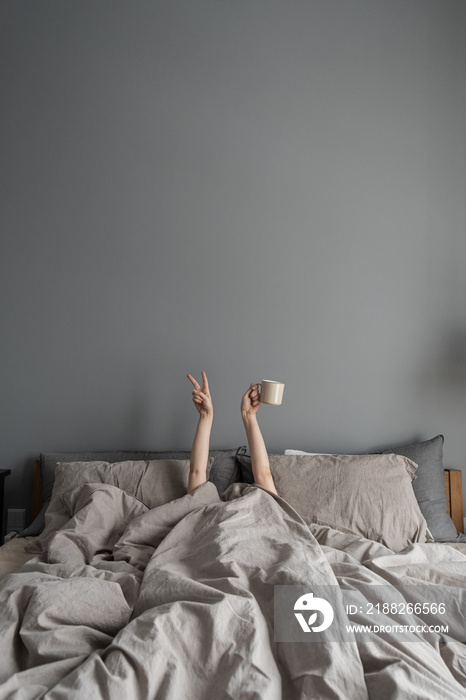 Woman laying in bed and holding mug with coffee with hand and show  Victory  symbol. Happy morning concept. Modern interior design with bedroom, bed, crumpled linen, pillows against grey wall.