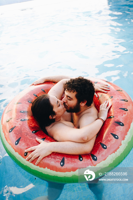 Young couple in love kissing and hugging on watermelon lilo in swimming pool at hotel resort. Summer vacation concept. Love moment.