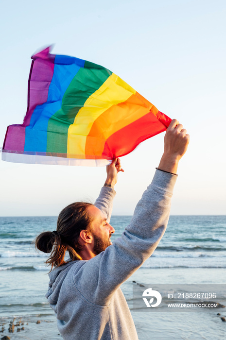 Silhouette of a man holding a gay pride rainbow flag blowing in the wind on a tropical beach with golden sunshine