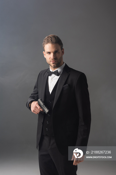 Confident businessman looking at camera while standing with gun on grey background with smoke