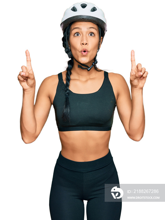 Beautiful hispanic woman wearing bike helmet amazed and surprised looking up and pointing with fingers and raised arms.