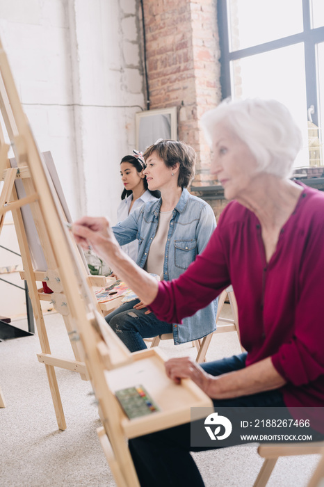 Portrait of art students sitting in row and painting at easels in art studio, focus on smiling  adult  woman enjoying work in center