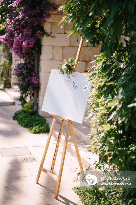 Blank wooden easel on the background of the wall and flowering bushes. Side view