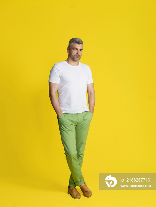 Handsome middle aged grey hair man posing with hands in pockets and legs crossed wearing white t-shirt and green jeans isolated on yellow background. Fit, muscular middle aged man