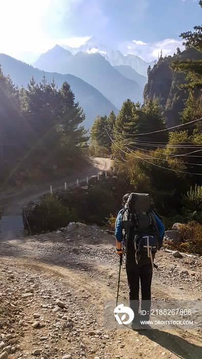 A man with a big black backpack hikes along a pathway on Annapurna Circuit Trek in Himalayas, Nepal. There are mountain chains around him. In the back there are massive barren mountain ranges