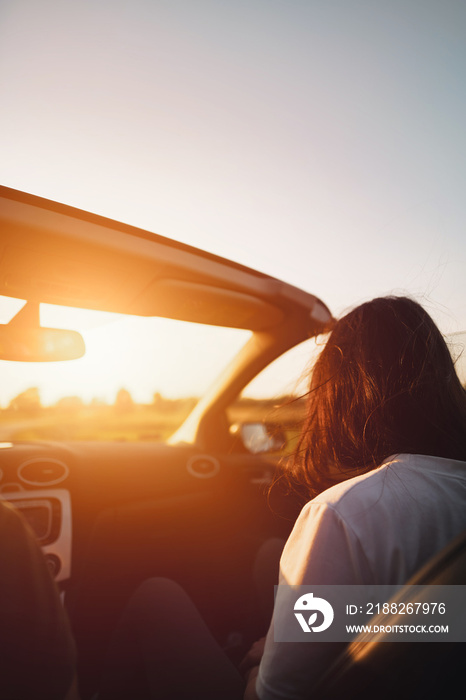 Rearview of an attractive brunette woman riding in the convertible car. Concept of summer adventures and new experiences. Strong sunlight during warm and long summer evenings.