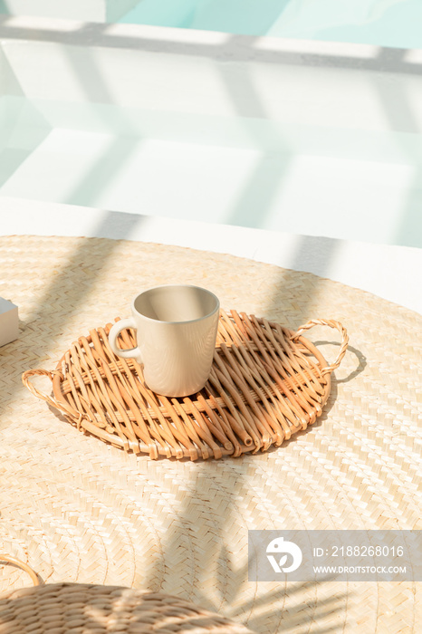 The mug is placed on a rattan tray. Swimming pool area. Decoration. Summer time. Vacation. Beach house. Relaxing space. Scandinavian style. Holiday vacation. Sunlight. Cozy space.