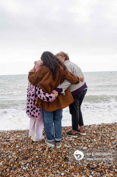 Grandmother, mother and daughter embracing on beach on cloudy day